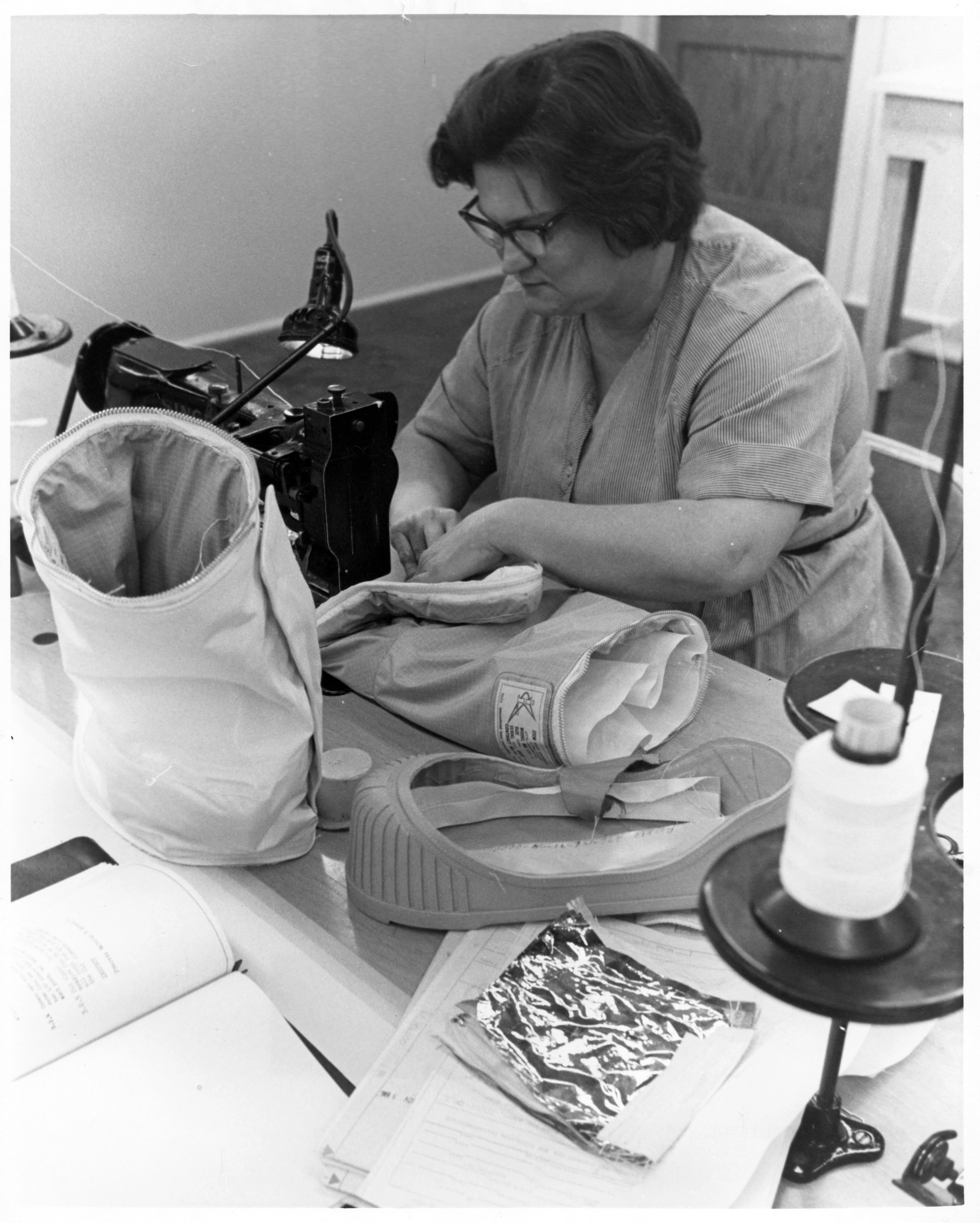 Black and white photo of a woman with short hair and black-framed glasses sitting at a sewing machine, assembling components of lunar overshoes.